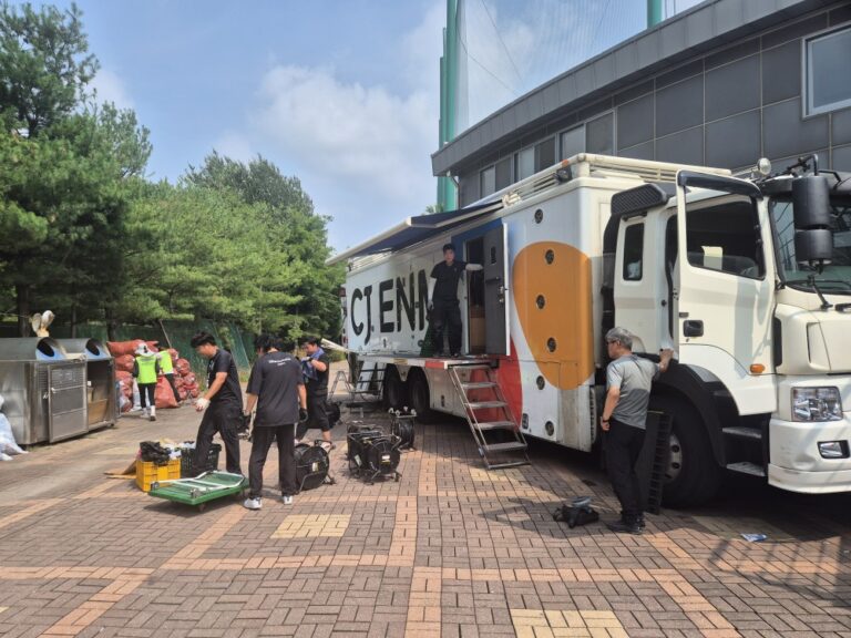야외 방송 제작 현장에 배치된 대형 방송 중계차와 전력 공급 가설 케이블 및 안전 점검 구역 전경